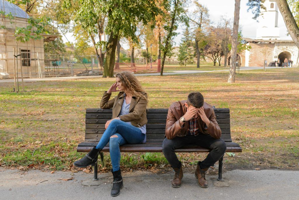 Infidelidad: Estrategias para Superarla y Fortalecer Tu Relación" – Imagen de una pareja sentada en un banco de parque con evidente tensión emocional. Ambos están distanciados físicamente y se cubren el rostro con las manos, reflejando angustia y desconexión. Ella mira hacia otro lado con expresión de enojo o tristeza, mientras él se inclina hacia adelante en una postura de frustración o arrepentimiento. El entorno es un parque con árboles y senderos, lo que añade un contraste entre la calma del lugar y la carga emocional de la escena. La imagen simboliza el impacto emocional de la infidelidad y la necesidad de sanar la relación.