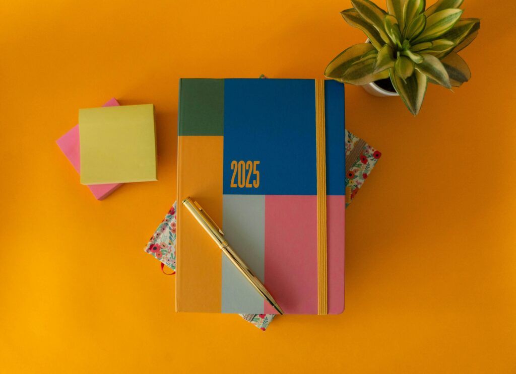Reflexiona sobre tus Propósitos 2025. Imagen de un cuaderno o agenda 2025 acompañada de coloridos post-its en tonos naranja, amarillo, rosado, azul y verde, junto a una planta similar a una sansevieria. El fondo naranja vibrante simboliza un 2025 lleno de oportunidades y energía positiva.